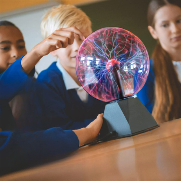 Lampe boule de plasma sur socle noir, posée sur un table marron, avec des enfant dans le fond un peu flou. Une jeune fille avance son bras vers la lampe et pose son doigt sur la boule de vere de la lampe plasma, et on voit le bras portant une manche longue bleu qui pose sa main sur le socle de la lampe. La lampe est allumée, on voit des faisceaux bleus et rouges.