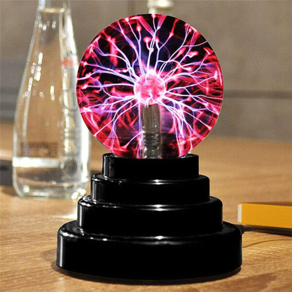 Lampe boule à plasma sur socle noir en escalier rond, allumée. On voit les faisceaux bleus et rouges. La lampe est posée sur une table en bois avec une bouteille d'eau flou dans le fond.